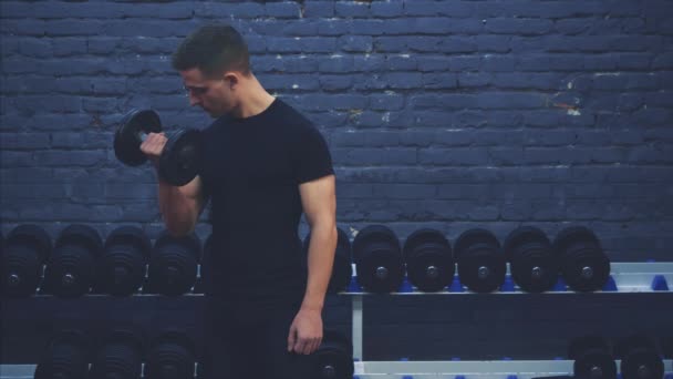 Un chico joven con una camiseta negra. Una gran barra en el gimnasio regularmente entrena las manos sobre un fondo oscuro. Durante esto, ella adora sus pezones . — Vídeo de stock