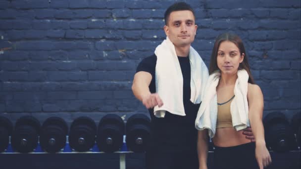 Young sporty couple guy and girl are in the gym. After a training session, they are hugging their fingers forward. During this, they have white towels. — Stock Video