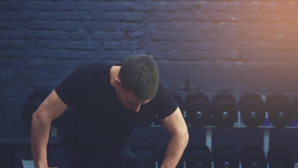 Ένα αθλητικό νεαρό αγόρι φορώντας ένα μαύρο μπλουζάκι ενώ καθόταν στο γυμναστήριο. Ένας άνθρωπος από ένα αθλητικό σώμα. Κατά τη διάρκεια αυτής της περιόδου αναπνέει ρυθμικά. Είναι στο γυμναστήριο.. — Αρχείο Βίντεο