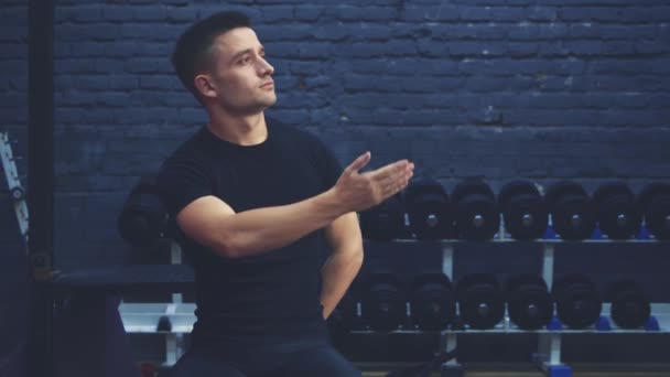 Un joven deportivo con una camiseta negra sentada en el gimnasio. Hombre con cuerpo deportivo. Durante este tiempo muestra su mano a un lado. Están en el gimnasio. . — Vídeos de Stock