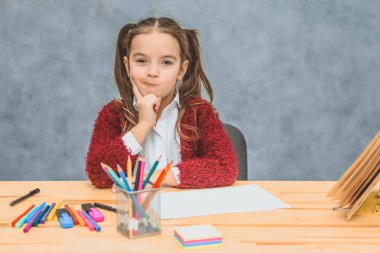 Gri bir arka plan üzerinde akıllı kız öğrenci. Elini çenesine koyarak hayalet gibi bir bakış attı. Masada gül renkli kalemler var..