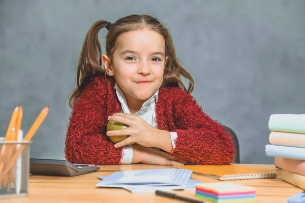 Mycket bra Schoolgirl flicka sitter vid bordet. Under detta håller han ett grönt äpple i handen och lägger en hand på handen. Leende. — Stockfoto