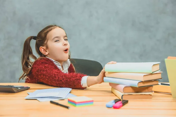 Très bonne écolière assise à la table. Pendant ce temps, il tient la main sur les livres qui se tiennent sur la table. En regardant la caméra a une expression faciale surprise . — Photo