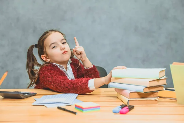 Very good schoolgirl girl sitting at the table. During this, he holds his hand on the books that stand on the table. Looking at the camera shows the index finger up. Has a surprised facial expression.