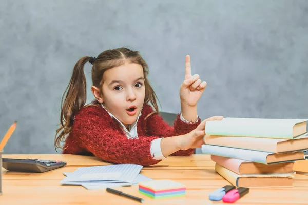 Très bonne écolière assise à la table. Pendant ce temps, il tient la main sur les livres qui se tiennent sur la table. En regardant la caméra montre l'index vers le haut. A une expression faciale surprise . — Photo