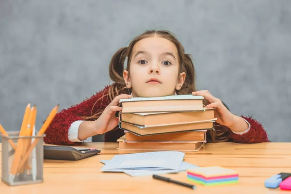 Ganska bra Schoolgirl flicka sitter vid bordet. Under detta, hålla huvudet på de böcker som står på bordet. Titta på kameran har en förvånad ansiktsuttryck. — Stockfoto