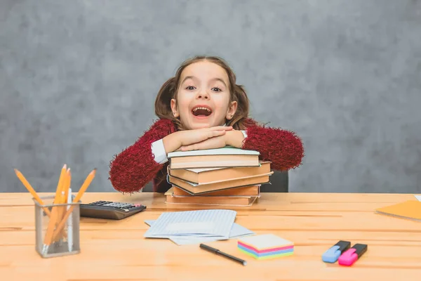 Ganska bra Schoolgirl flicka sitter vid bordet. Under detta, hålla huvudet på de böcker som står på bordet. Att sätta en hand på handen tittar på kameran öppnar munnen. — Stockfoto