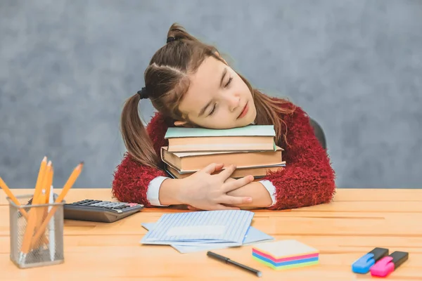 Kinder- und Schulbedarf auf dem Hintergrund einer grauen Wand. Selbstgemachte Aufgabe und Rückkehr zum Schulkonzept. Eine Schülerin sitzt auf ihrem Schreibtisch mit buntem Papeterie und Schulbüchern. das Mädchen schläft auf einem Bündel — Stockfoto