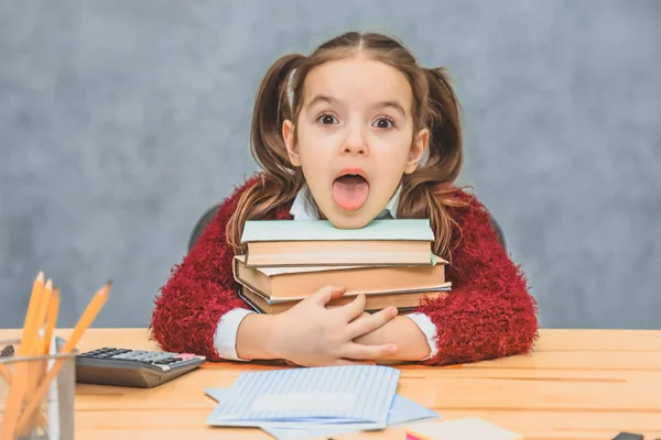 En lekfull söt liten flicka är att ha kul samtidigt förlita sig på tjocka böcker på en grå bakgrund. Hennes hår är tillverkat i selar. — Stockfoto