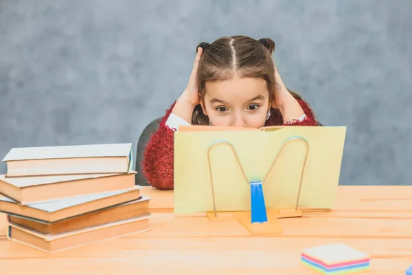 Милая школьница на сером фоне. Работает домашнее задание, читая книгу в желтой обложке. От удивления она положила его руки на голову . — стоковое фото