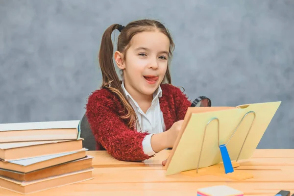 Studentessa intelligente si siede a tavola su uno sfondo grigio. Durante questo, legge il libro con una lente d'ingrandimento. Mostrando una lingua guarda la fotocamera . — Foto Stock