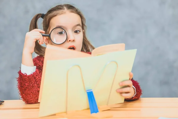 Une écolière intelligente s'assoit à la table sur un fond gris. Pendant ce temps, elle lit le livre avec une loupe. Regarde attentivement le livre, jaune . — Photo