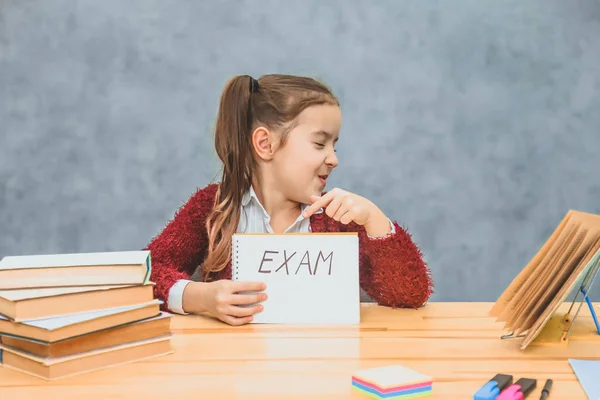 Una bella scolaretta è triste e sopraffatta. Documento di conduzione con esame di testo. Isolato su uno sfondo grigio. Mostra l'indice sulla parola esame girando la testa di lato . — Foto Stock