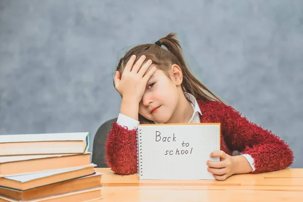 Een klein schoolmeisje op een grijze achtergrond. Houd het papier met de tekst terug naar school. Nadat hij zijn hoofd naar beneden liet, zette het meisje haar hand op haar hoofd. — Stockfoto