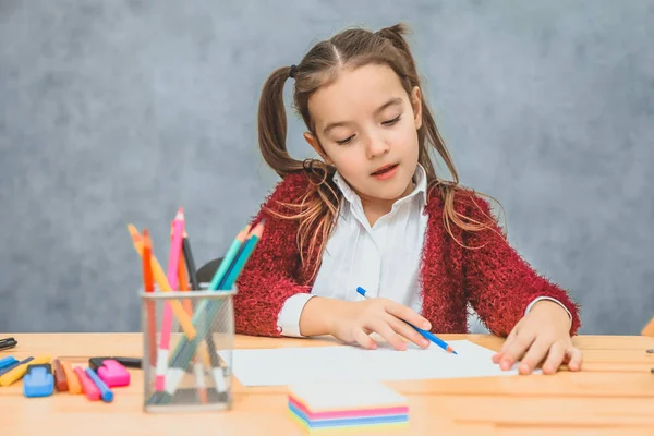 Bella ragazza su sfondo grigio. È impegnato a disegnare una matita blu. Sul tavolo ci sono matite, pennarelli, foglie . — Foto Stock