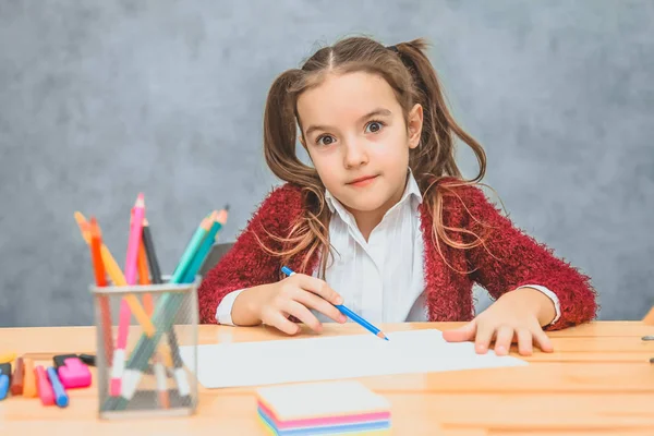 Bella ragazza su sfondo grigio. Prendendo una matita blu guarda nella fotocamera. Sul tavolo ci sono matite di colore rosa . — Foto Stock
