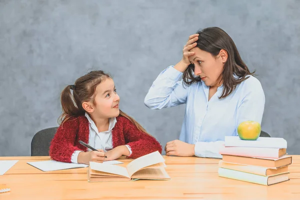Genç anne ve kızı gri bir arka plan üzerinde oturuyor. Bu esnada anne, bir kızın ev ödevini nasıl yaptığına dikkat eder. Sürprizden elini alnına koyar.. — Stok fotoğraf