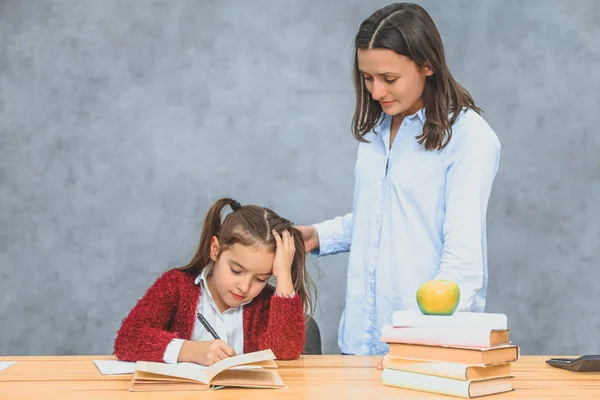 Moeder en dochter op een grijze achtergrond. Op hetzelfde moment schrijft de school jongen huiswerk, moeder horloges haar. — Stockfoto