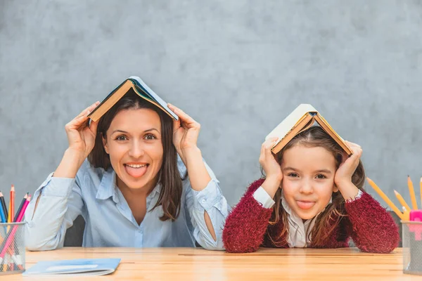 Mãe e filha alegres em um fundo cinza. Durante isso, na cabeça de minha mãe e filha, há livros abertos. Olhando para a câmera mostrar a língua . — Fotografia de Stock