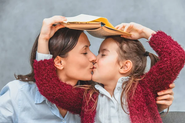 Mamá y su hija sobre un fondo gris. Abrazo de besos y colegiala sosteniendo la cabeza de las madres y su libro abierto. De cerca. . — Foto de Stock