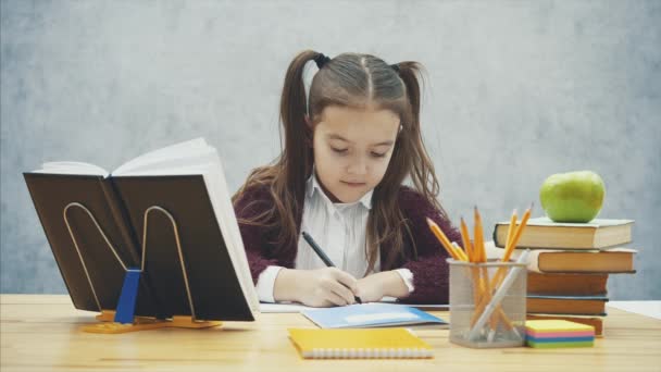 회색 배경에 지능형 여학생입니다. 이 시간 동안 그는 테이블에 앉아있다. 숙제를 쓴다. — 비디오