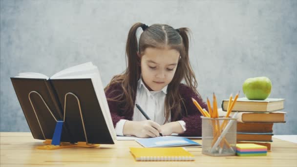 Colegiala inteligente sobre un fondo gris. Durante este tiempo está sentado a la mesa. Escribe la tarea . — Vídeo de stock