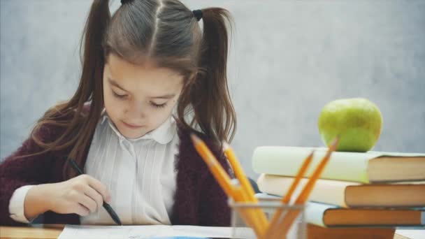 Ein Schulmädchen auf grauem Hintergrund. währenddessen sitzt er am Tisch und nimmt ein Notizbuch in die Hand. — Stockvideo