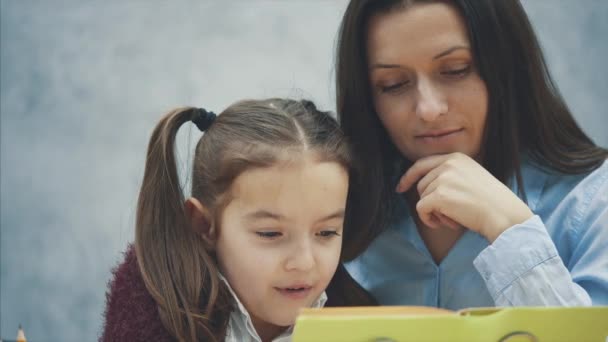 Maman et fille s'assoient à la table et lisent un livre. Pendant ce fond gris . — Video