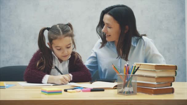 Mutter und Tochter sitzen am Tisch vor grauem Hintergrund. Hausaufgaben schreiben, kommunizieren. — Stockvideo