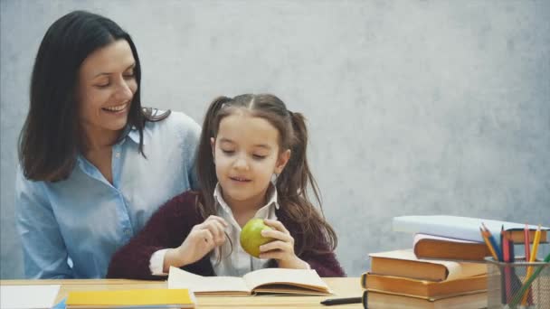 Una hija diligente lee un libro. Mamá trajo una manzana verde. Siéntate sobre un fondo gris . — Vídeos de Stock