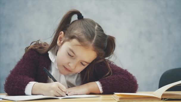 Sumienny uczennica pisze pracę domową. W tym czasie siedzi przy stole na szarym tle. Po upuszczeniu głową w dół. — Wideo stockowe
