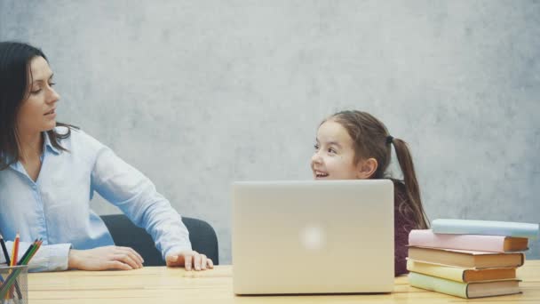 Een schoolmeisje zit aan tafel. Tijdens deze, het kost tijd voor een laptop. Gelukkig. Op een grijze achtergrond. — Stockvideo