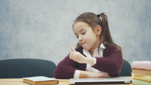 Una estudiante está sentada en la mesa. Durante este tiempo, elige un libro o una computadora portátil. Feliz En un fondo gris . — Vídeo de stock