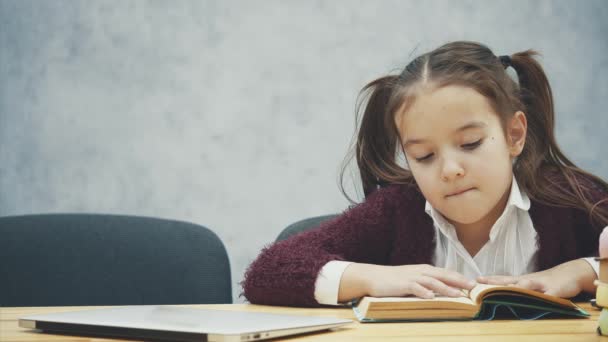 Une écolière est assise à la table. Pendant ce temps, il lit un livre. La fermeture du livre prend l'ordinateur portable. Heureux. Sur un fond gris . — Video