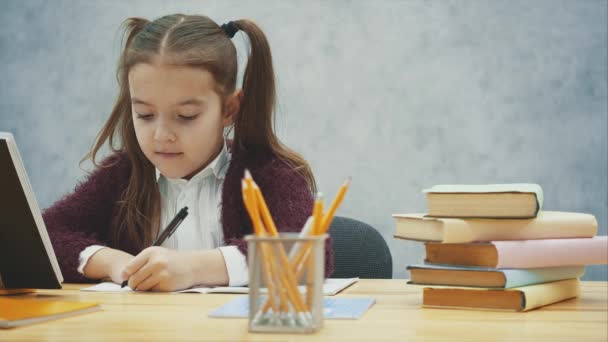 Una buena colegiala se sienta a la mesa sobre un fondo gris. Durante esto, escribe los deberes mientras mira el libro. Mociones lentas . — Vídeo de stock