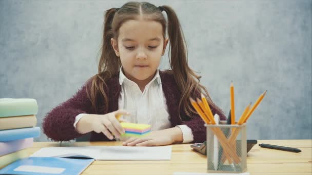 Μια καλή μαθήτρια κάθεται στο τραπέζι σε γκρι φόντο. Κατά τη διάρκεια αυτού, ζωγραφίζει ένα χαμόγελο σε πολύχρωμα φύλλα και χαμόγελα. — Αρχείο Βίντεο
