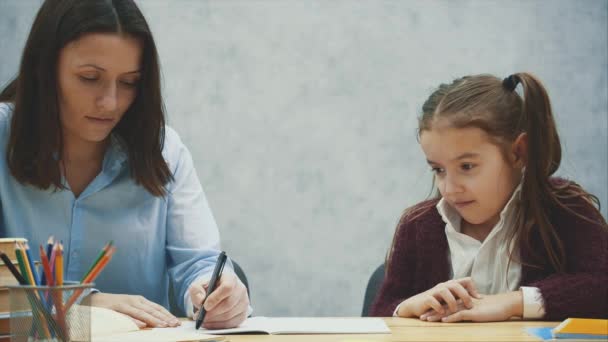 灰色の背景にママと娘をクローズアップ。この間、彼女は笑顔の娘のために宿題を書きます。関係の概念. — ストック動画
