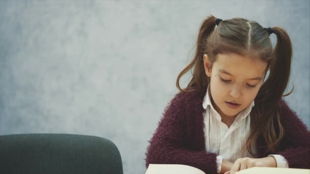 特写女婴看书坐在灰色的室内。女学生正在学习课本。校服的孩子乐于读书。家庭培训一本有趣的儿童读物。慢 — 图库视频影像