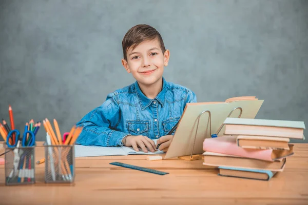 Young School Kid i blå jeans t-shirt sitter vid bordet med böcker, skriva, titta på kameran, leende. — Stockfoto