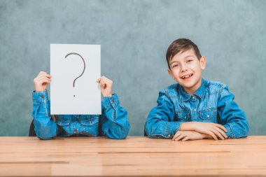 Masabaşında oturan öğrenciler. Kız ünlem işareti olan kağıdın arkasına saklanıyor. Çocuk kameraya gülümsüyor..