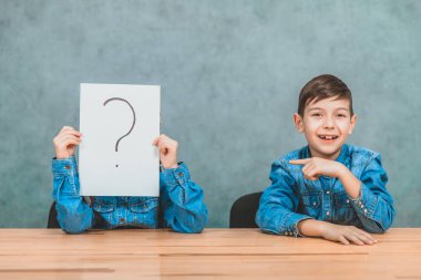 Masabaşında oturan öğrenciler. Kız ünlem işareti olan kağıdın arkasına saklanıyor. Çocuk onu parmağıyla işaret ediyor, gülümsüyor..