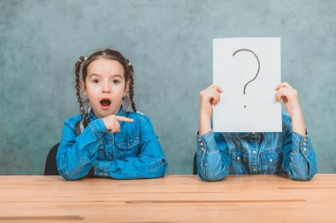 Masabaşında oturan öğrenciler. Çocuk ünlem işareti olan kağıdın arkasına saklanıyor. Komik pigtails ile kız, onu parmağıyla işaret ediyor, asaletli bakıyor.