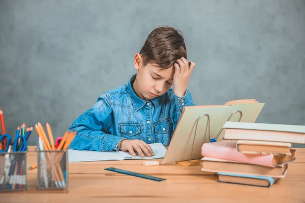 Pequeno estudante cansado está sentado na mesa, sobrecarregado com livros, segurando a mão na testa . — Fotografia de Stock
