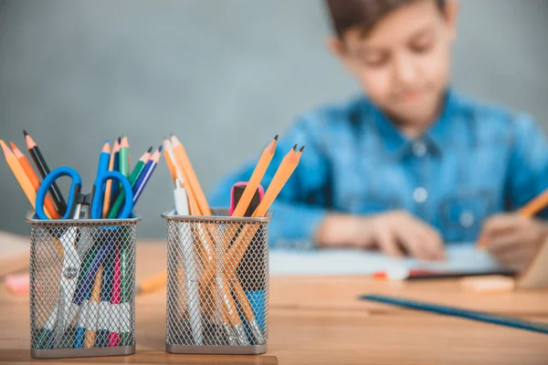 Två korgar med färgade blyertspennor, saxar och överstrykningspennor i framkant. Schoolboy skriver på suddig bakgrund. — Stockfoto