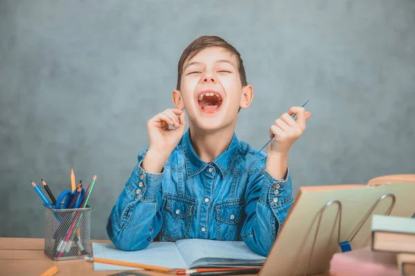 Rolig skolpojke sitter vid skrivbordet full av skolmaterial, skrattar. — Stockfoto