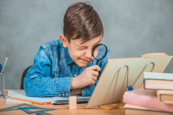 Nyfiken liten pojke tittar genom förstoringsglas på något i sin bok som detektiv. — Stockfoto