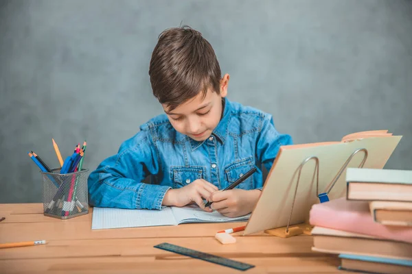 Jovem escola concentrada criança em jeans azul t-shirt escrita sentado à mesa com livros . — Fotografia de Stock