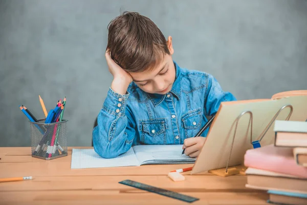 Młody skoncentrowany dziecko szkoły w niebieskie dżinsy t-shirt piśmie siedzi przy stole pełnym książek. — Zdjęcie stockowe