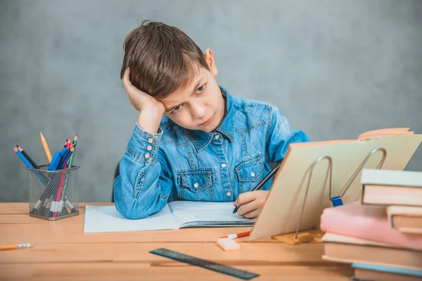 Weinig vermoeide school jongen zit aan de balie, overweldigd door boeken, leunend zijn hoofd op de hand. — Stockfoto