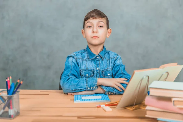 Smart allvarlig skolpojke sitter med självsäker ansiktsuttryck, hans händer viks på skrivbordet. — Stockfoto